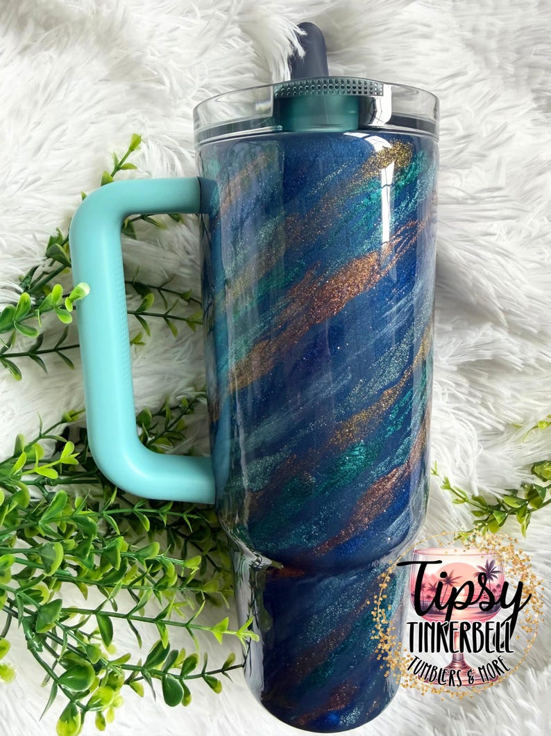 Puede incluir: Un vaso grande y aislado con un asa y tapa de color turquesa. El vaso presenta un dise&ntilde;o arremolinado de purpurina azul, turquesa y dorada. Las palabras "Tipsy Tinkerbell Tumblers & More" est&aacute;n impresas en la parte inferior.