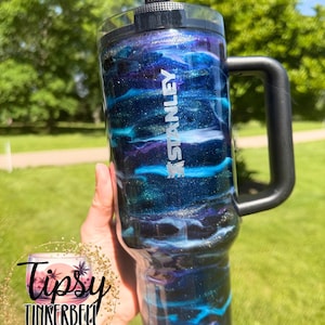 May include: A Stanley tumbler with a black handle and lid. The tumbler features a swirling design of blue, purple, and silver glitter, with the Stanley logo in white. The tumbler is held against a background of green grass and trees.