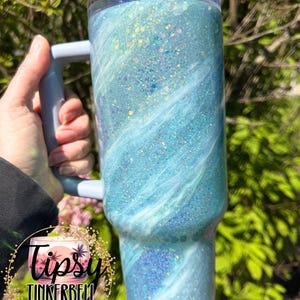May include: A large, glittery tumbler with a light blue and white swirl design. The tumbler has a handle and a clear lid. The text "Tipsy Tinkerbell Tumblers & More" is on the bottom left of the tumbler.