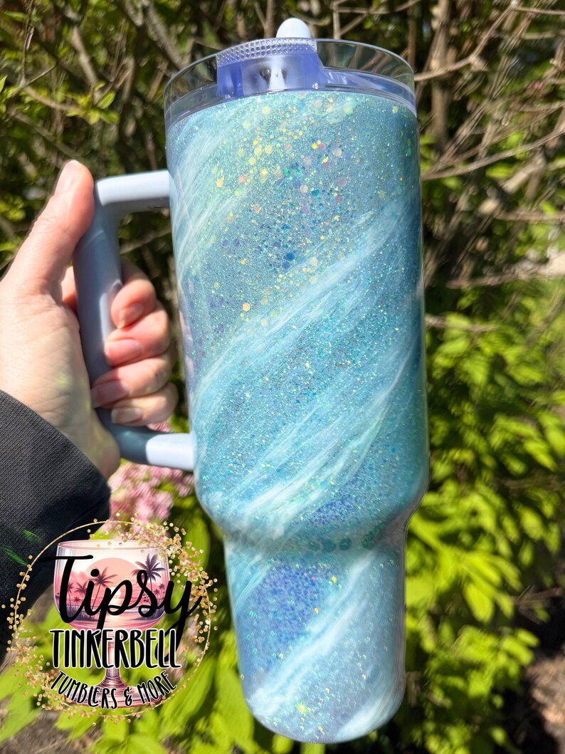 May include: A large, glittery tumbler with a light blue and white swirl design. The tumbler has a handle and a clear lid. The text "Tipsy Tinkerbell Tumblers & More" is on the bottom left of the tumbler.