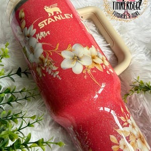 K&ouml;nnte beinhalten: Ein roter Stanley-Becher mit Glitzer und einem Blumenmuster aus wei&szlig;en und cremefarbenen Bl&uuml;ten. Der Becher hat einen beigen Griff und einen durchsichtigen Deckel. Das Stanley-Logo ist oben in Wei&szlig; aufgedruckt. Ein kreisf&ouml;rmiges Logo zeigt Tipsy Tinkerbell.