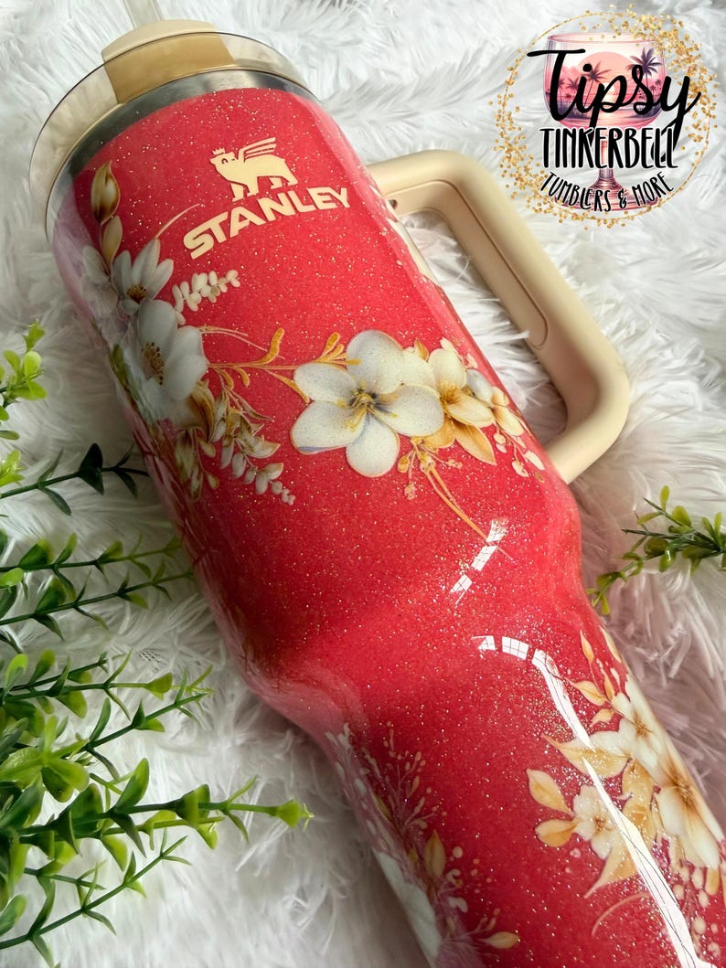 K&ouml;nnte beinhalten: Ein roter Stanley-Becher mit Glitzer und einem Blumenmuster aus wei&szlig;en und cremefarbenen Bl&uuml;ten. Der Becher hat einen beigen Griff und einen durchsichtigen Deckel. Das Stanley-Logo ist oben in Wei&szlig; aufgedruckt. Ein kreisf&ouml;rmiges Logo zeigt Tipsy Tinkerbell.