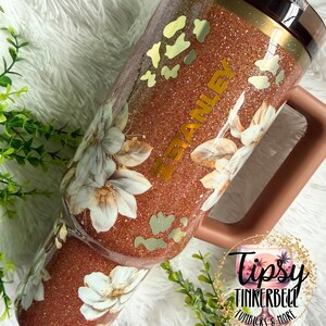 May include: A copper-coloured glitter tumbler with a handle and a dark lid. The tumbler features a floral pattern with white flowers and gold-toned leopard print accents. The word STANLEY is printed vertically in gold lettering on the side of the cup.