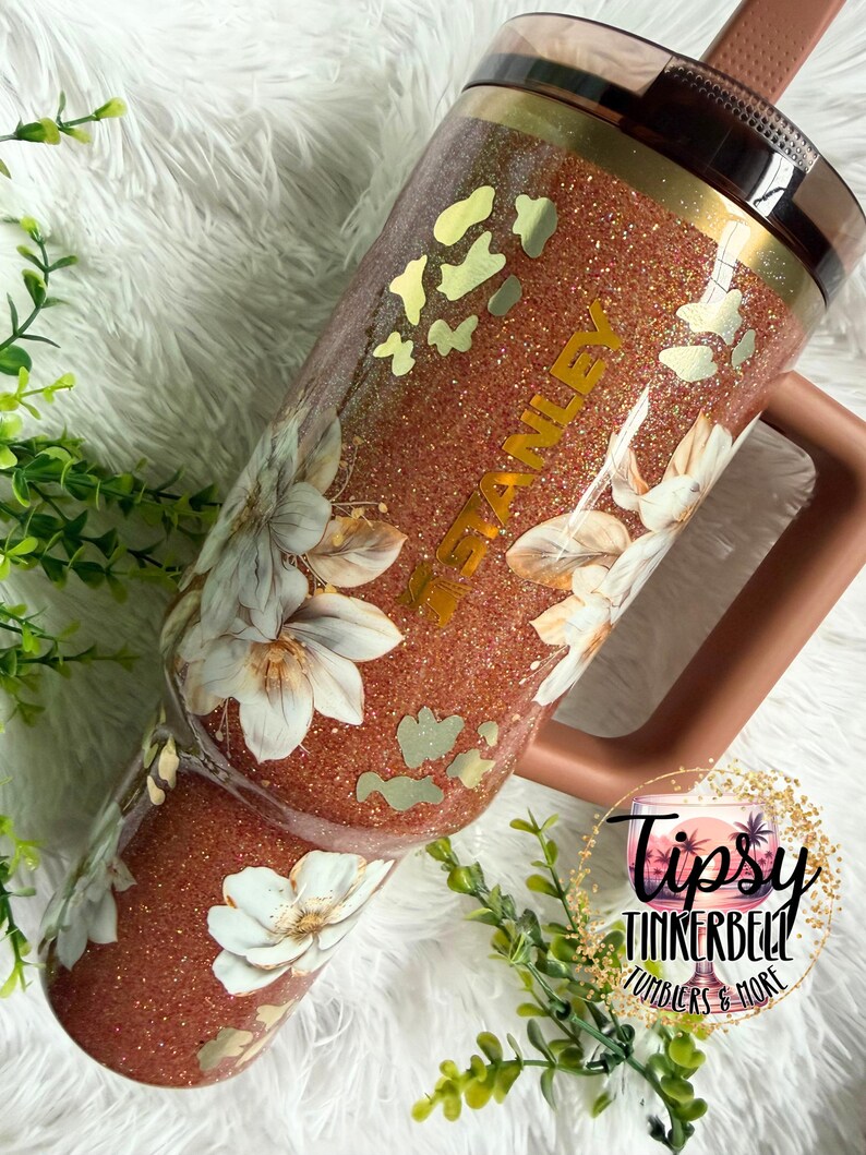 May include: A copper-coloured glitter tumbler with a handle and a dark lid. The tumbler features a floral pattern with white flowers and gold-toned leopard print accents. The word STANLEY is printed vertically in gold lettering on the side of the cup.