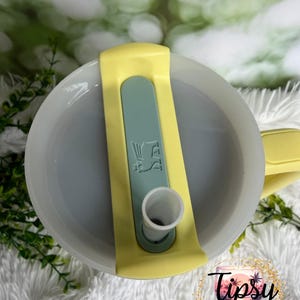 May include: A close-up view of a tumbler lid. The lid is light gray with a yellow handle and a light green slider. The slider has a white spout and a graphic. The bottom right corner has a logo that says "Tipsy Tinkerbell Tumblers & More."