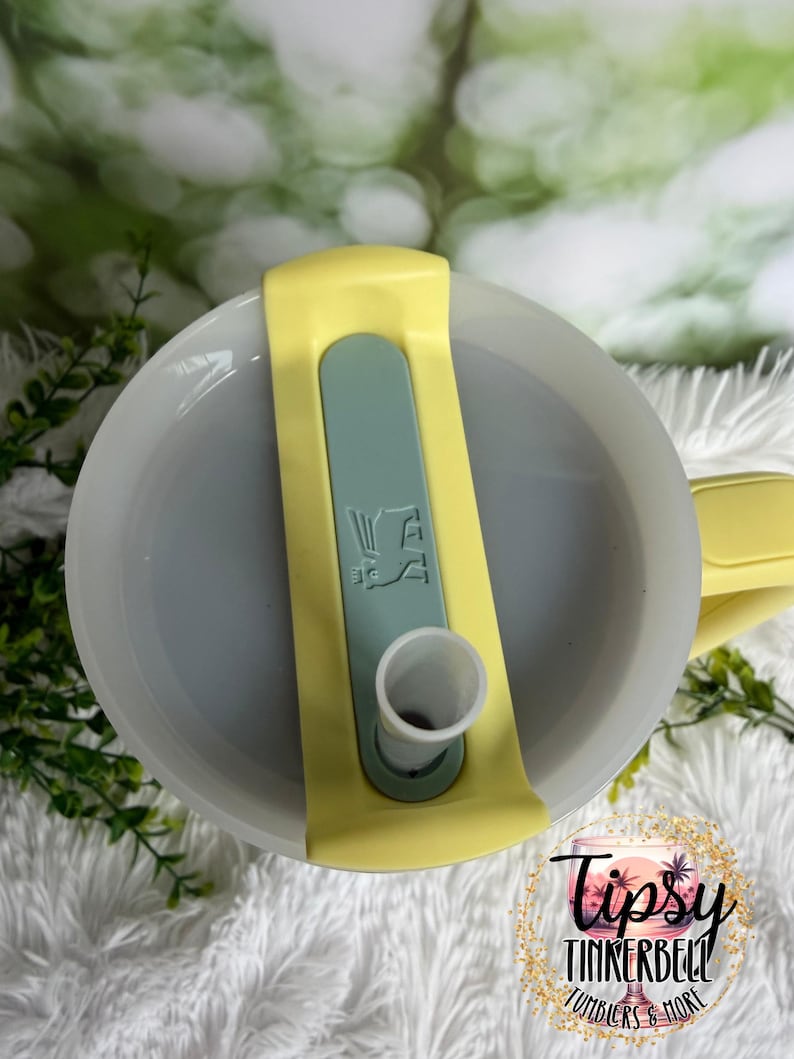 May include: A close-up view of a tumbler lid. The lid is light gray with a yellow handle and a light green slider. The slider has a white spout and a graphic. The bottom right corner has a logo that says "Tipsy Tinkerbell Tumblers & More."