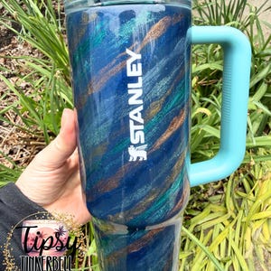 Puede incluir: Un vaso grande y aislado con un dise&ntilde;o de remolino azul, verde azulado y dorado. El vaso tiene un asa azul claro y el logotipo "STANLEY" en blanco. El vaso est&aacute; dise&ntilde;ado para mantener las bebidas fr&iacute;as o calientes. El vaso mide aproximadamente 23 cm de alto.