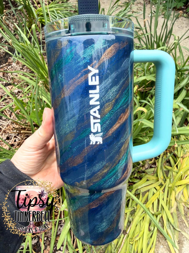 Puede incluir: Un vaso grande y aislado con un dise&ntilde;o de remolino azul, verde azulado y dorado. El vaso tiene un asa azul claro y el logotipo "STANLEY" en blanco. El vaso est&aacute; dise&ntilde;ado para mantener las bebidas fr&iacute;as o calientes. El vaso mide aproximadamente 23 cm de alto.