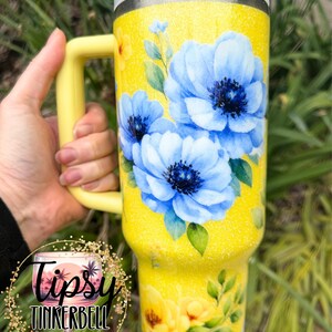 May include: A yellow glitter tumbler with a handle, featuring a floral design of blue and yellow flowers with green leaves. The tumbler has a silver rim and a yellow lid. The text "Tipsy Tinkerbell Tumblers & More" is visible.