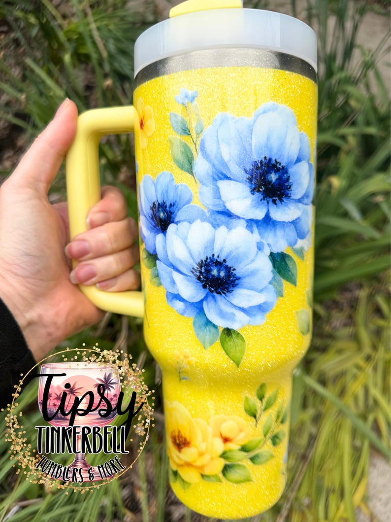 May include: A yellow glitter tumbler with a handle, featuring a floral design of blue and yellow flowers with green leaves. The tumbler has a silver rim and a yellow lid. The text "Tipsy Tinkerbell Tumblers & More" is visible.
