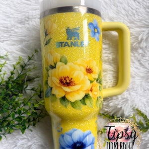 May include: A yellow glitter Stanley tumbler with a handle, featuring floral designs in yellow and blue. The tumbler has the Stanley logo and a straw. The text "Tipsy Tinkerbell Tumblers & More" is also visible.