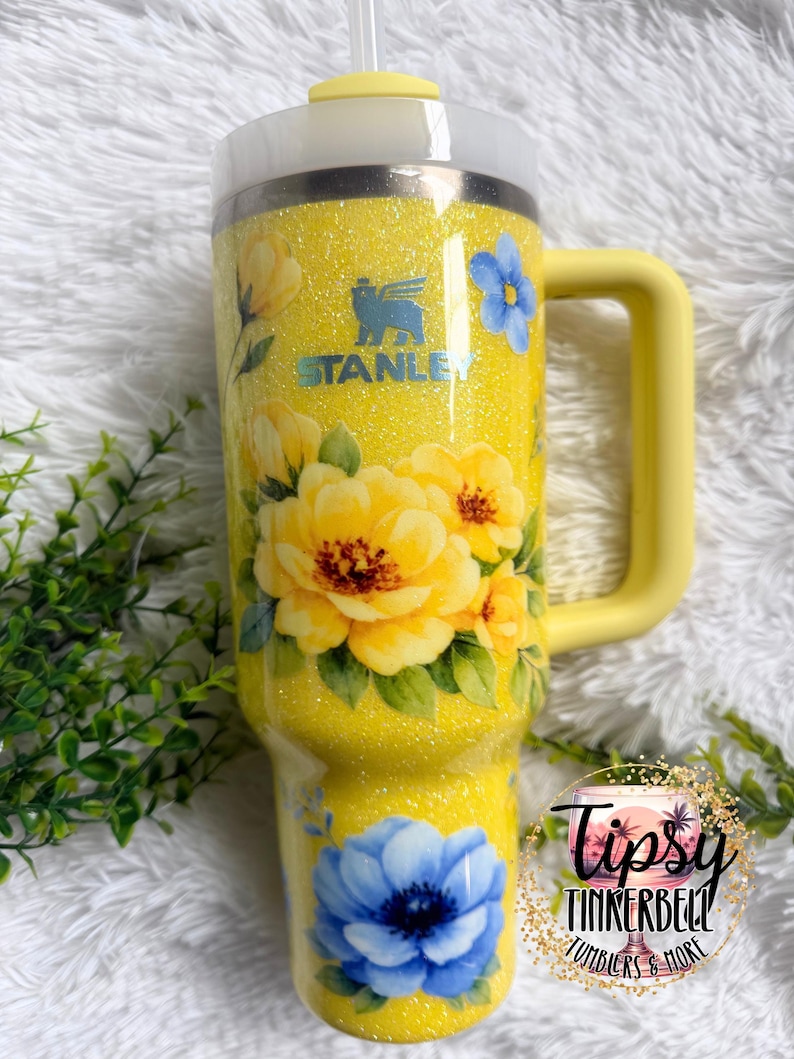 May include: A yellow glitter Stanley tumbler with a handle, featuring floral designs in yellow and blue. The tumbler has the Stanley logo and a straw. The text "Tipsy Tinkerbell Tumblers & More" is also visible.