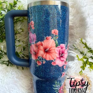 Może przedstawiać: Ciemnoniebieski kubek z brokatem, uchwytem i przezroczystą pokrywką, ozdobiony kwiatowym wzorem r&oacute;żowych i pomarańczowych kwiat&oacute;w hibiskusa. W prawym dolnym rogu obrazu widoczne jest okrągłe logo z napisem Tipsy Tinkerbell Tumblers & More.