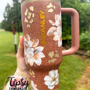 May include: A copper-coloured glitter tumbler with a handle and a lid, featuring the word STANLEY vertically in gold lettering. The surface is decorated with white floral illustrations and gold leopard print spots scattered across the textured finish.
