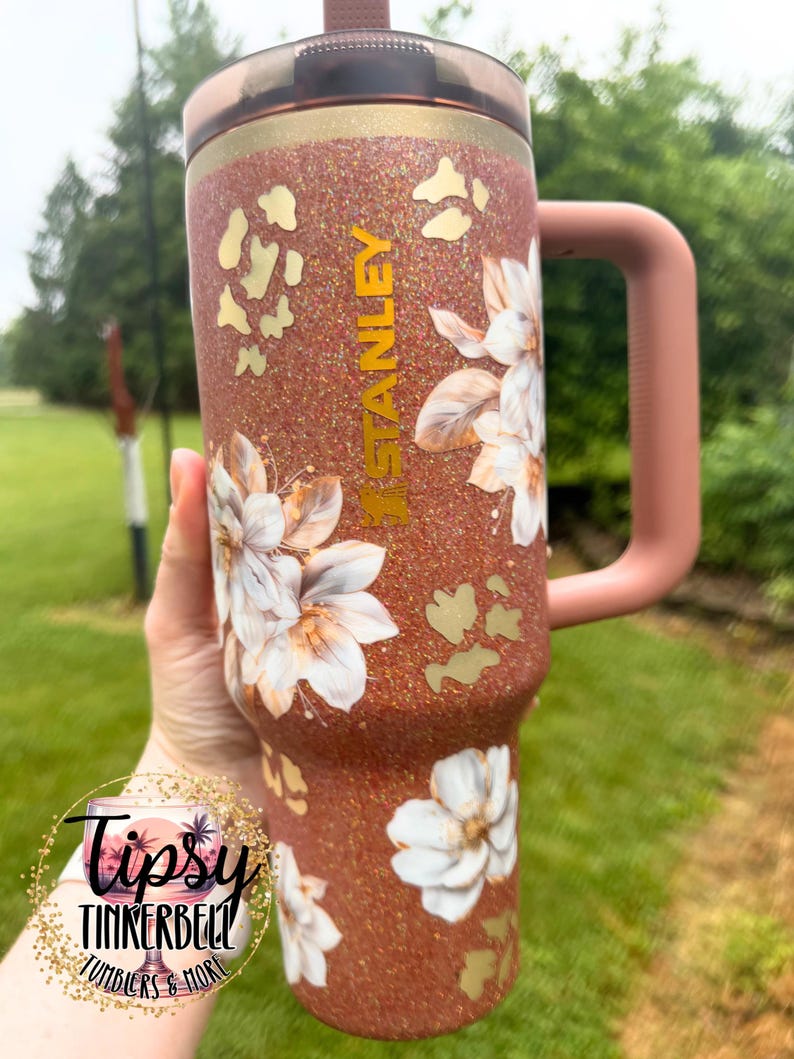 May include: A copper-coloured glitter tumbler with a handle and a lid, featuring the word STANLEY vertically in gold lettering. The surface is decorated with white floral illustrations and gold leopard print spots scattered across the textured finish.