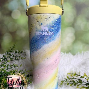 May include: A Stanley tumbler with a yellow handle and lid. The body of the tumbler features a swirling design of blue, yellow, pink, and white with glitter accents. The Stanley logo is visible on the front. The tumbler is a pastel color scheme.