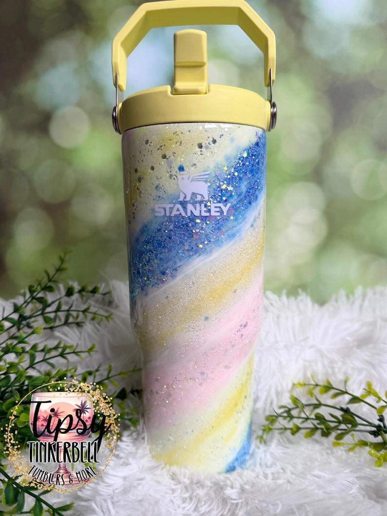 May include: A Stanley tumbler with a yellow handle and lid. The body of the tumbler features a swirling design of blue, yellow, pink, and white with glitter accents. The Stanley logo is visible on the front. The tumbler is a pastel color scheme.