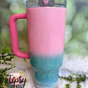 May include: A stainless steel tumbler with a pink handle and a clear lid featuring a pink straw. The body has a glittery pink top section that fades into a teal glitter middle, transitioning to a white and sandy beige base at the bottom.