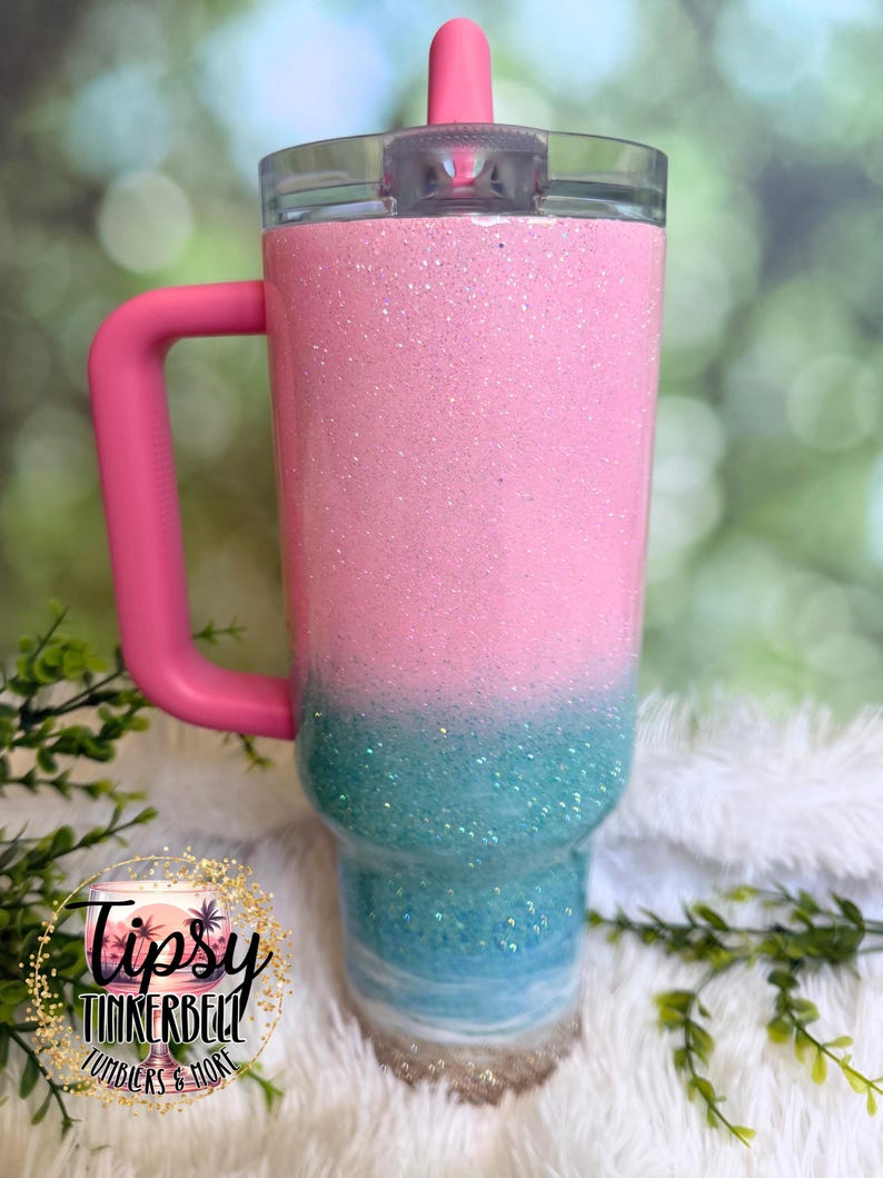 May include: A stainless steel tumbler with a pink handle and a clear lid featuring a pink straw. The body has a glittery pink top section that fades into a teal glitter middle, transitioning to a white and sandy beige base at the bottom.