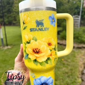 May include: Yellow glitter Stanley tumbler with a floral design. The tumbler features large yellow and blue flowers, a yellow handle, and a white lid. The Stanley logo is visible. The tumbler has a straw.