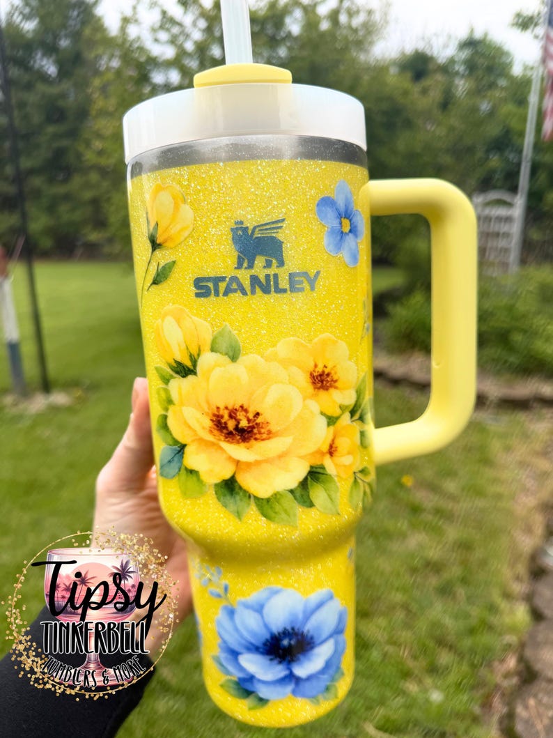 May include: Yellow glitter Stanley tumbler with a floral design. The tumbler features large yellow and blue flowers, a yellow handle, and a white lid. The Stanley logo is visible. The tumbler has a straw.