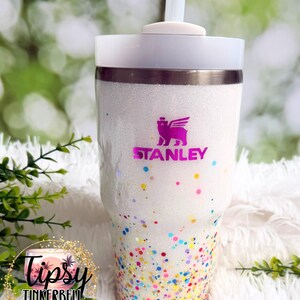 Puede incluir: Un vaso blanco con purpurina, tapa y pajita. El vaso presenta un logotipo rosa de Stanley y detalles coloridos en forma de confeti. La parte inferior del vaso est&aacute; cubierta de puntos de colores. El vaso es de Tipsy Tinkerbell Tumblers & More.