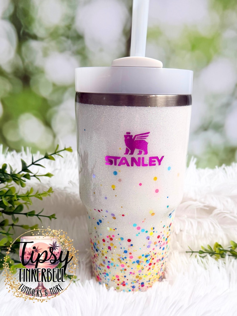 Puede incluir: Un vaso blanco con purpurina, tapa y pajita. El vaso presenta un logotipo rosa de Stanley y detalles coloridos en forma de confeti. La parte inferior del vaso est&aacute; cubierta de puntos de colores. El vaso es de Tipsy Tinkerbell Tumblers & More.
