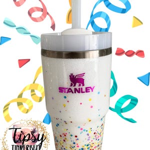 Puede incluir: Vaso blanco brillante con tapa y pajita blancas. El vaso presenta el logotipo "Stanley" en morado y est&aacute; decorado con confeti colorido. La parte inferior del vaso tiene un dise&ntilde;o de confeti. Tambi&eacute;n es visible el logotipo de "Tipsy Tinkerbell Tumblers & More".