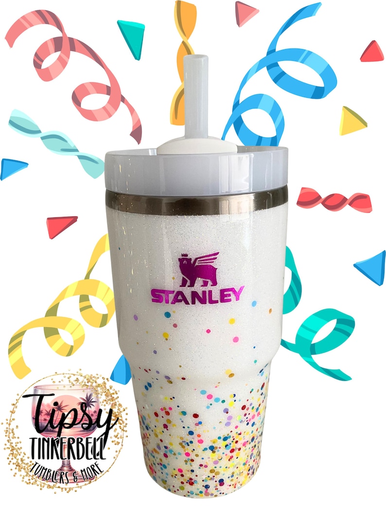 Puede incluir: Vaso blanco brillante con tapa y pajita blancas. El vaso presenta el logotipo "Stanley" en morado y est&aacute; decorado con confeti colorido. La parte inferior del vaso tiene un dise&ntilde;o de confeti. Tambi&eacute;n es visible el logotipo de "Tipsy Tinkerbell Tumblers & More".