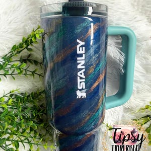Puede incluir: Un vaso Stanley azul y verde azulado con asa y tapa verde azulado. El vaso tiene un dise&ntilde;o brillante con la palabra "STANLEY" en blanco. El vaso est&aacute; decorado con detalles en verde y dorado. Tiene una tapa transparente y una pajita negra.