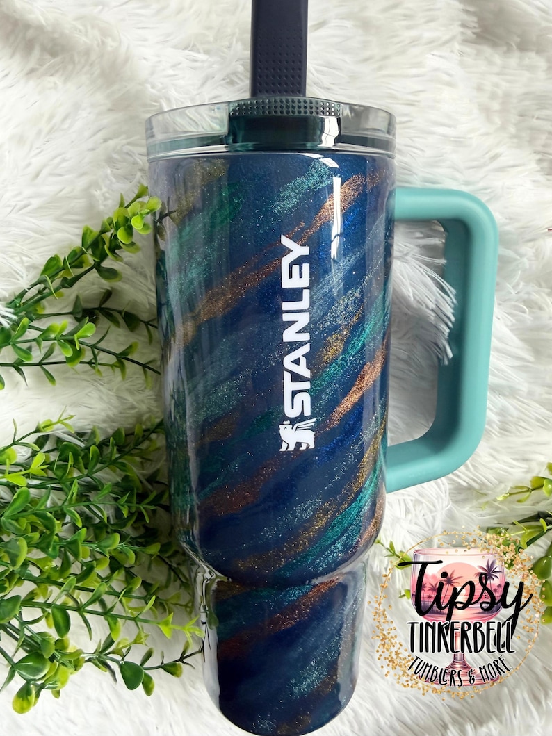 Puede incluir: Un vaso Stanley azul y verde azulado con asa y tapa verde azulado. El vaso tiene un dise&ntilde;o brillante con la palabra "STANLEY" en blanco. El vaso est&aacute; decorado con detalles en verde y dorado. Tiene una tapa transparente y una pajita negra.