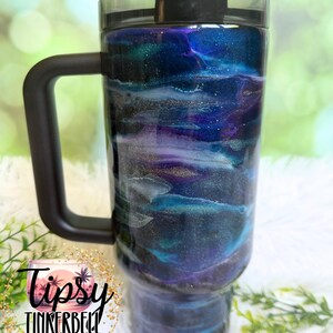 May include: A large tumbler with a black handle and lid. The tumbler features a swirling design in shades of blue, purple, and white, with gold accents. The text "Tipsy Tinkerbell Tumblers & More" is visible on the side.