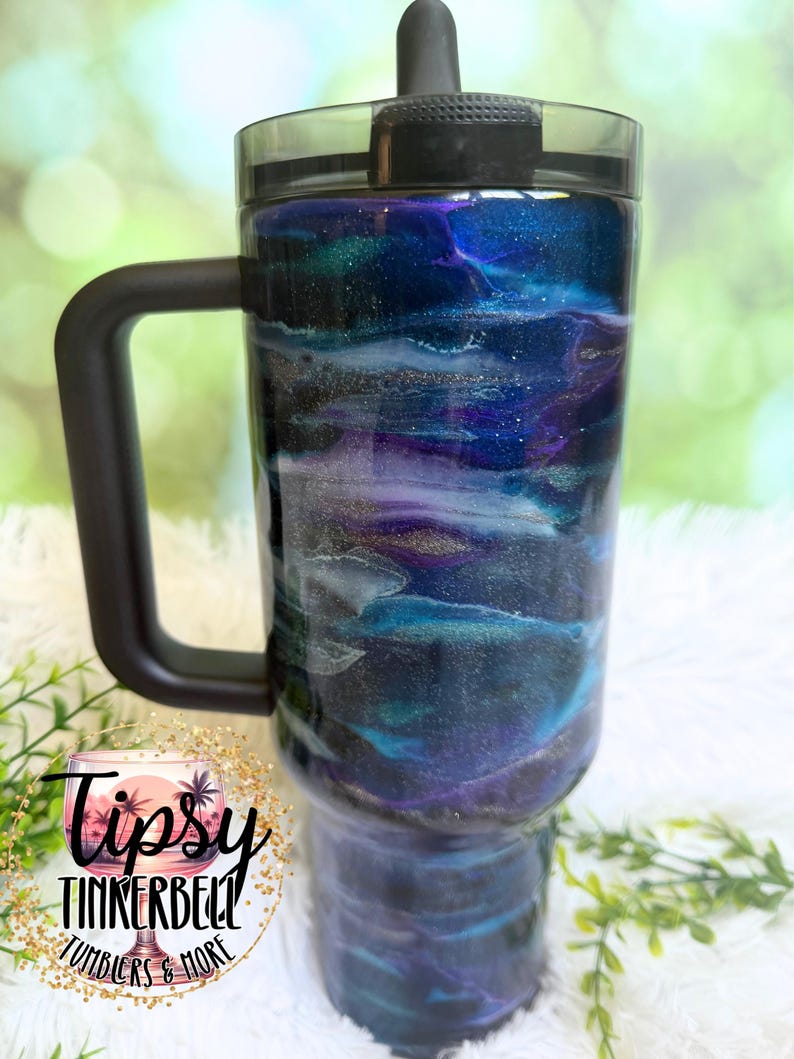 May include: A large tumbler with a black handle and lid. The tumbler features a swirling design in shades of blue, purple, and white, with gold accents. The text "Tipsy Tinkerbell Tumblers & More" is visible on the side.