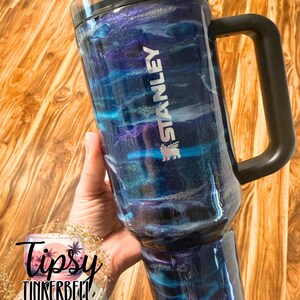 May include: A Stanley tumbler with a black handle and lid, featuring a swirling blue, purple, and silver design. The tumbler has the Stanley logo and the text "Tipsy Tinkerbell Tumblers & More" is visible.