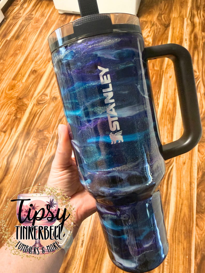 May include: A Stanley tumbler with a black handle and lid, featuring a swirling blue, purple, and silver design. The tumbler has the Stanley logo and the text "Tipsy Tinkerbell Tumblers & More" is visible.