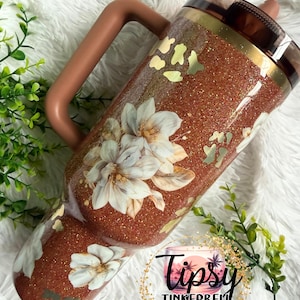 May include: A copper-coloured glitter tumbler with a handle, featuring white floral illustrations and gold leopard print accents. The tumbler has a dark brown lid and a straw. A circular logo with the text Tipsy TINKERBELL TUMBLERS & MORE is visible.