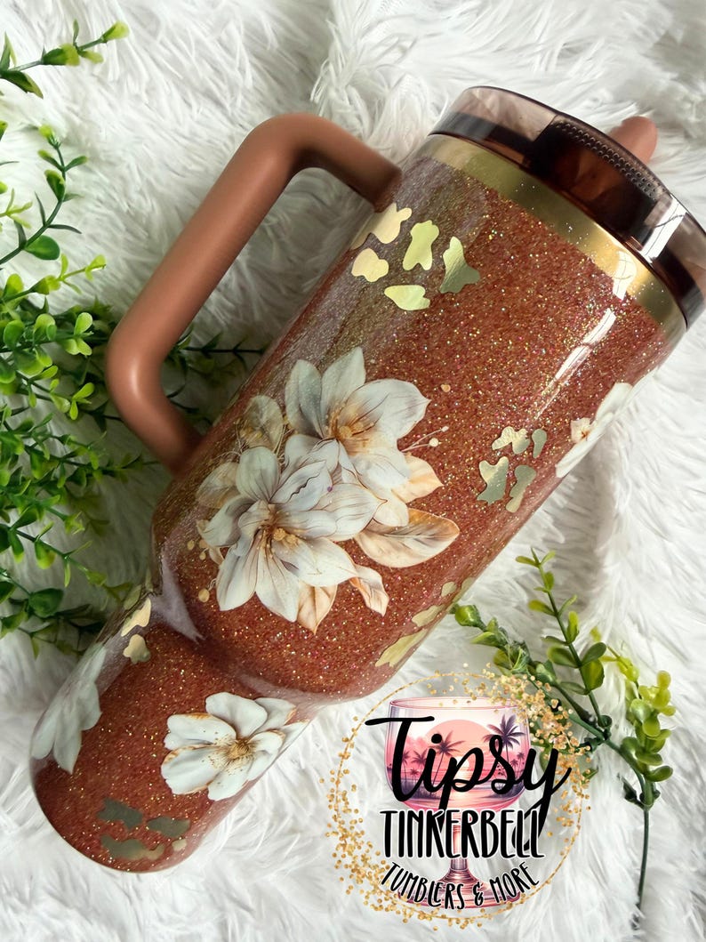 May include: A copper-coloured glitter tumbler with a handle, featuring white floral illustrations and gold leopard print accents. The tumbler has a dark brown lid and a straw. A circular logo with the text Tipsy TINKERBELL TUMBLERS & MORE is visible.