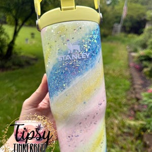 May include: A Stanley tumbler with a yellow handle and lid. The body of the tumbler is decorated with a swirling pattern of blue, yellow, and pink glitter. The Stanley logo is visible on the front. The tumbler is a hydration accessory.