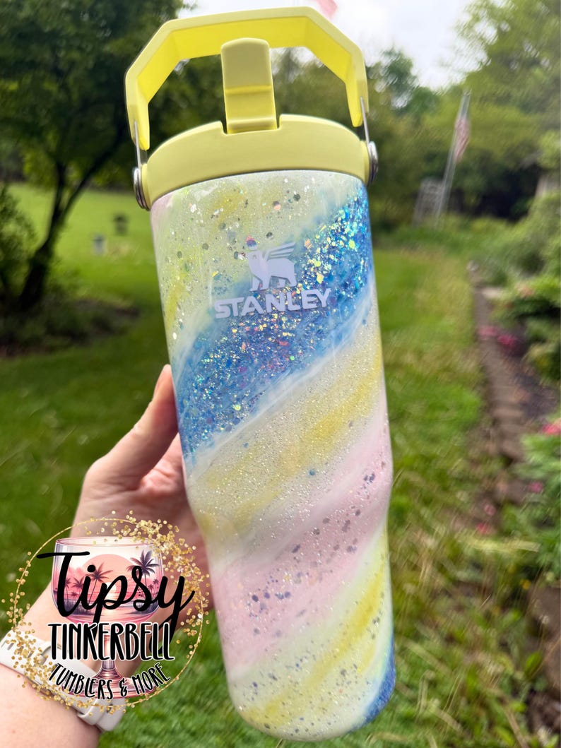 May include: A Stanley tumbler with a yellow handle and lid. The body of the tumbler is decorated with a swirling pattern of blue, yellow, and pink glitter. The Stanley logo is visible on the front. The tumbler is a hydration accessory.