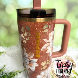 May include: A copper-coloured glitter tumbler with a handle, a dark translucent lid, and a vertical straw. The cup features a floral pattern, gold leopard print accents, and the word STANLEY printed vertically in gold on the front surface.
