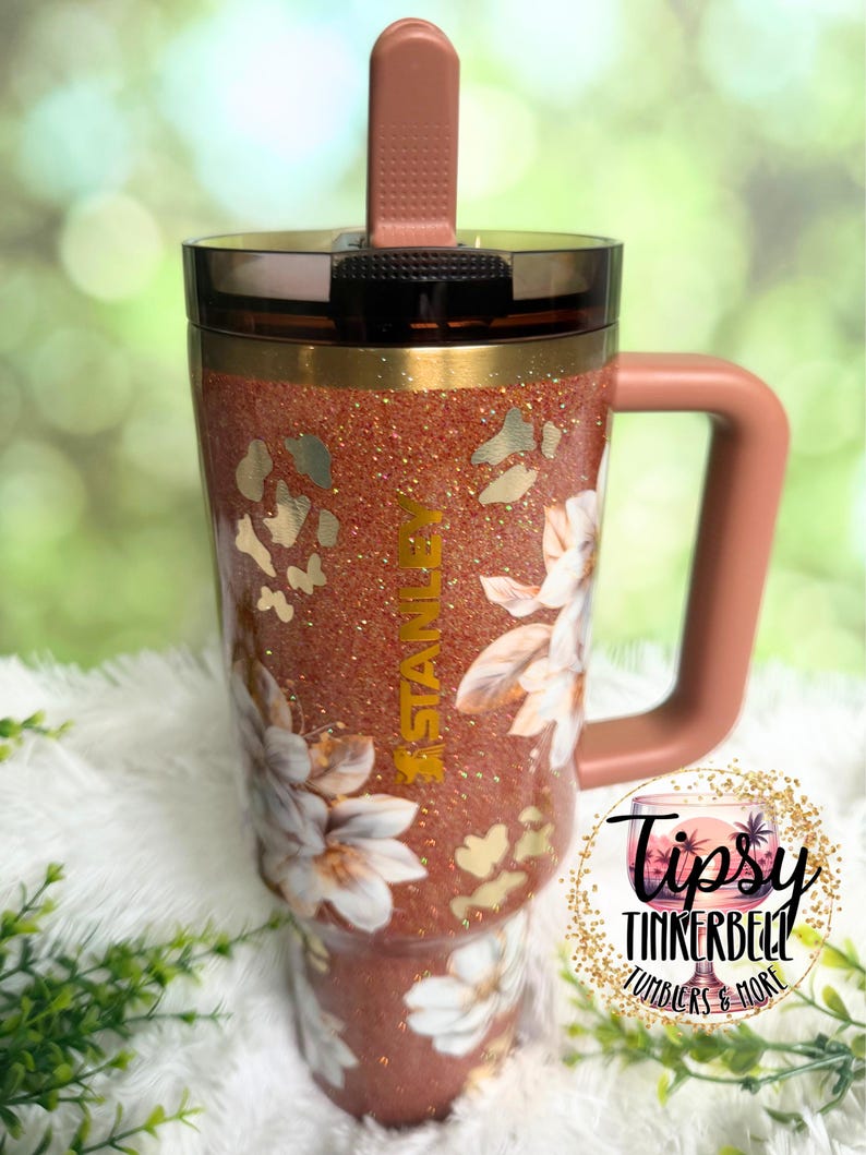 May include: A copper-coloured glitter tumbler with a handle, a dark translucent lid, and a vertical straw. The cup features a floral pattern, gold leopard print accents, and the word STANLEY printed vertically in gold on the front surface.
