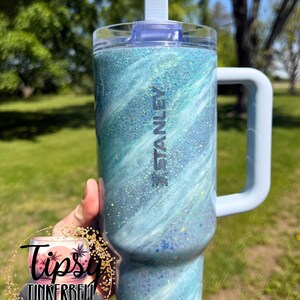 May include: A Stanley tumbler with a light blue and white glitter swirl design. The tumbler has a handle, a clear lid, and a white flip-top. The word "STANLEY" is printed on the side.