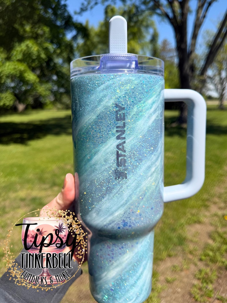May include: A Stanley tumbler with a light blue and white glitter swirl design. The tumbler has a handle, a clear lid, and a white flip-top. The word "STANLEY" is printed on the side.