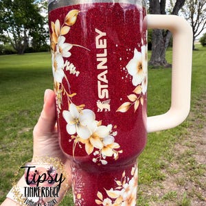 Puede incluir: Un vaso Stanley rojo brillante con un dise&ntilde;o floral. El vaso tiene una tapa transparente y un asa color crema. La palabra "STANLEY" est&aacute; impresa en blanco en el lateral. El vaso est&aacute; decorado con flores blancas y amarillas.