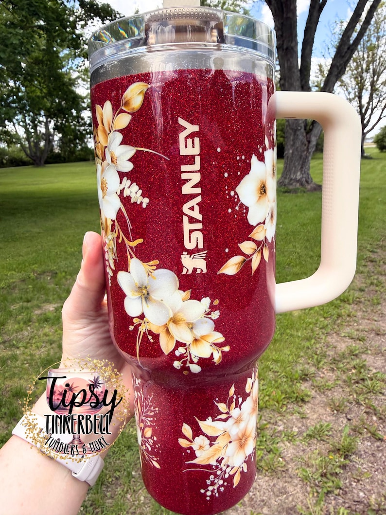Puede incluir: Un vaso Stanley rojo brillante con un dise&ntilde;o floral. El vaso tiene una tapa transparente y un asa color crema. La palabra "STANLEY" est&aacute; impresa en blanco en el lateral. El vaso est&aacute; decorado con flores blancas y amarillas.