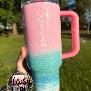 May include: A Stanley tumbler featuring a pink glitter top that transitions into a turquoise and blue ocean wave design, ending in a sandy-colored base. The cup includes a pink handle, a clear lid, and a pink straw, with the brand name STANLEY printed.