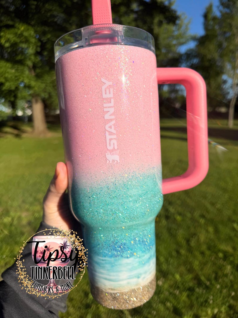 May include: A Stanley tumbler featuring a pink glitter top that transitions into a turquoise and blue ocean wave design, ending in a sandy-colored base. The cup includes a pink handle, a clear lid, and a pink straw, with the brand name STANLEY printed.