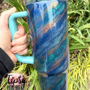 Puede incluir: Un vaso grande y aislado con un asa y tapa turquesa. El cuerpo del vaso est&aacute; pintado con un dise&ntilde;o arremolinado de azul, turquesa y dorado. El vaso se sostiene frente a un fondo de follaje verde.
