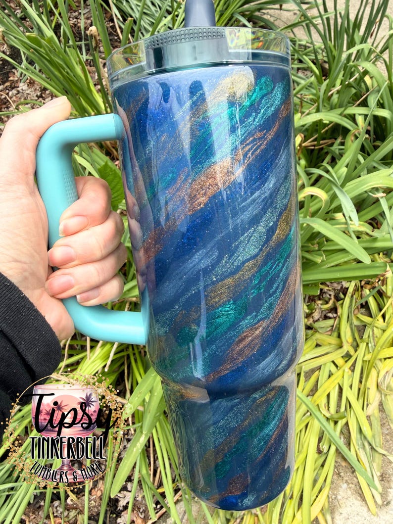 Puede incluir: Un vaso grande y aislado con un asa y tapa turquesa. El cuerpo del vaso est&aacute; pintado con un dise&ntilde;o arremolinado de azul, turquesa y dorado. El vaso se sostiene frente a un fondo de follaje verde.