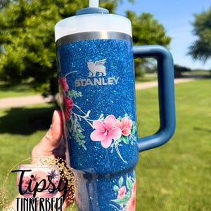 Może przedstawiać: Niebieski kubek Stanley z brokatowym wykończeniem, ozdobiony kwiatowym wzorem z r&oacute;żowymi kwiatami typu hibiskus i zielonymi liśćmi. Kubek posiada ciemnoniebieski uchwyt, białą pokrywkę i przezroczystą słomkę. Logo Stanley jest widoczne w kolorze srebrnym.